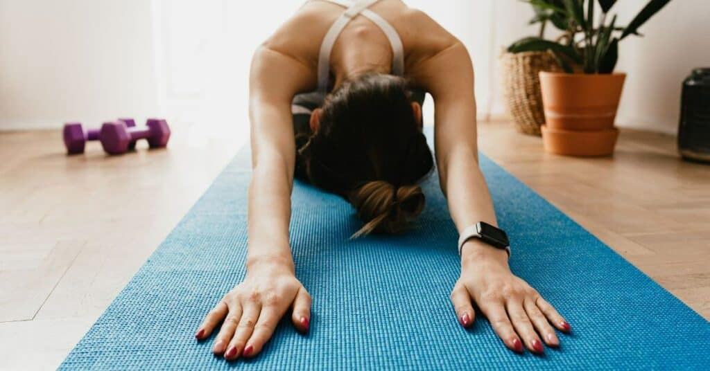 A thin woman with manicured nails reaches forward on her yoga mat in a child's pose, free weights and house plants in the background