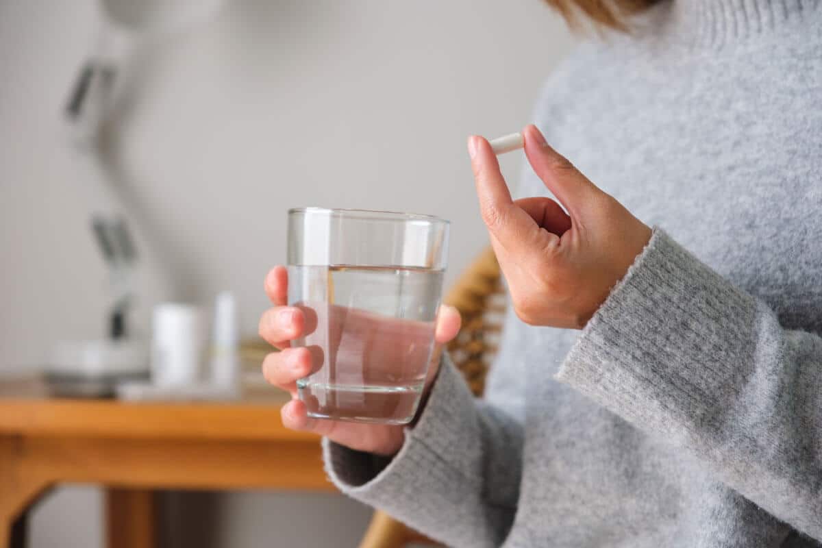 A woman in a knit sweater holds a glass of water in one hand a white pill capsule in the other, When’s the Best Time To Take Magnesium?