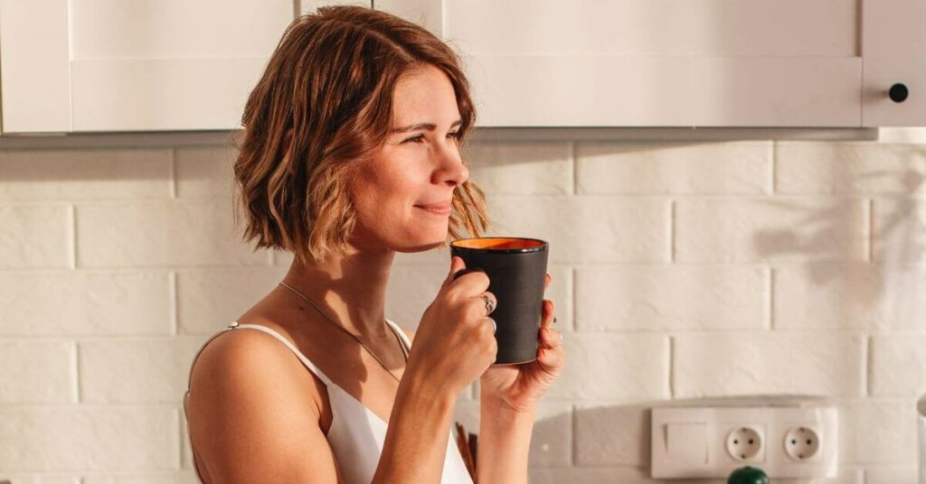 A woman looks off into the distance confidently and happily as she sips her morning coffee in a sunlit kitchen, Benefits of Magnesium Lysinate Glycinate