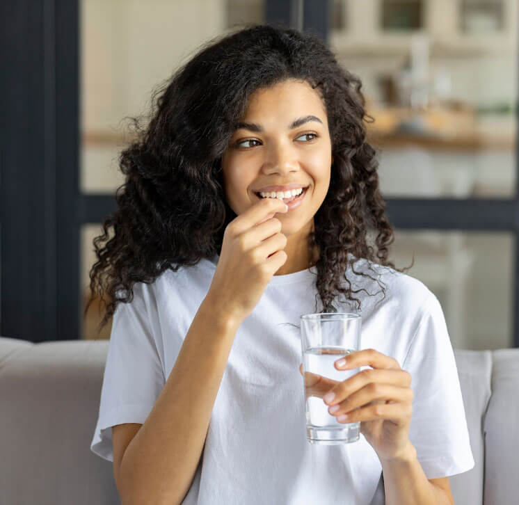 Young woman with curly hair and a glass of water in her hand smiles as she puts a More Chill magnesium and l-theanine capsule in her mouth